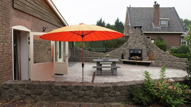 A patio area with a stone wall, a wooden bench, and a red umbrella. - Holiday Home in Olst-wijhe With Sauna (Olst)