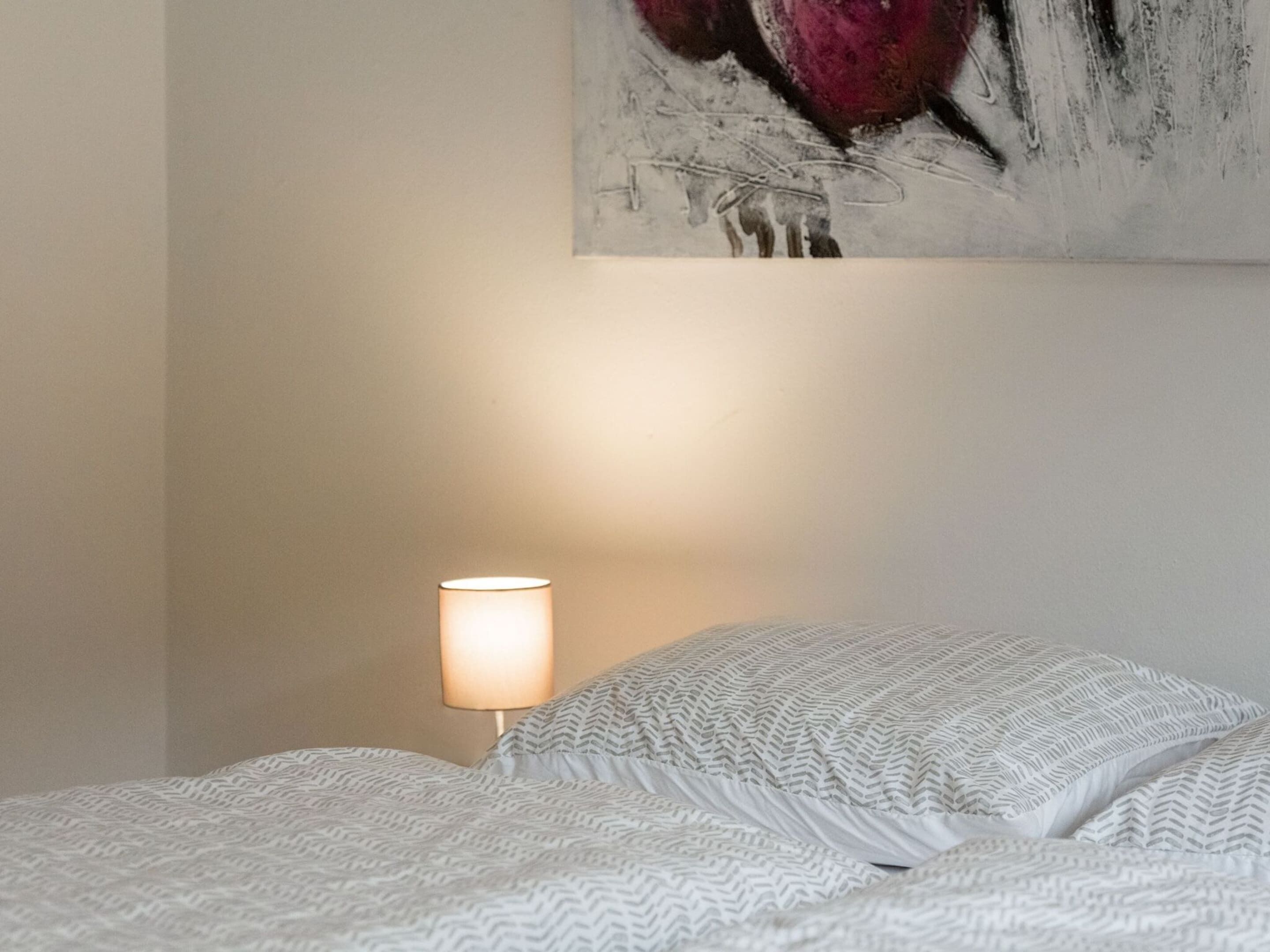 Een opgemaakt bed met behangpatroon, een bedlampje en een schilderij aan de muur.