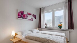 Ein Schlafzimmer mit zwei Betten, einem Nachttisch mit Lampe, einem Fenster mit Vorhängen und Blick nach draußen.