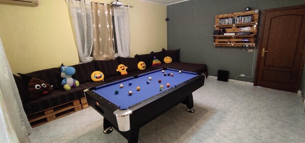 Une table de billard dans une pièce où se trouvent un canapé et des coussins, une bibliothèque et une porte.