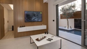 Un salon moderne avec un téléviseur à écran plat fixé sur un mur en bois, une table basse blanche avec des télécommandes, et une vue sur une piscine à l’extérieur.