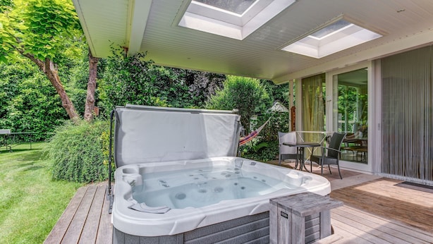 Un bain à remous sur une terrasse en bois, avec un patio couvert, le tout entouré de verdure.