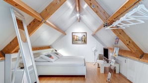 Une chambre à coucher aménagée dans un grenier, avec un plafond en pente, des poutres en bois, un lit, un bureau et un radiateur.