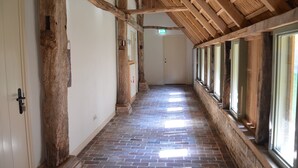 A hallway with exposed wooden beams, brick flooring, and large windows along one side. - Farmhouse in Geesteren Near Forests (Geesteren)