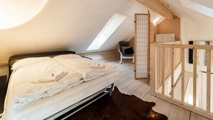 Un dormitorio con cama, una silla, piso de madera y techo inclinado con tragaluz.