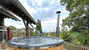 Ein hölzernes Whirlpool auf einer Terrasse mit Metallgeländer, umgeben von Bäumen und einem bewölkten Himmel.