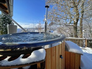 A hot tub with a wooden deck, surrounded by snow-covered trees and a clear sky.