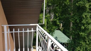 A balcony with a brown awning and white railing, overlooking a green landscape with trees and a building with a green roof. - Enjoy the nature on Zlatibor (Zlatibor)