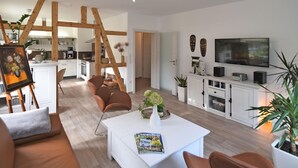 A modern living room with a white sofa, a coffee table, and a TV. There is a kitchen in the background with wooden beams. - Ferienwohnung mit Sauna in Schonbrunn Thuringen (Schleusegrund)