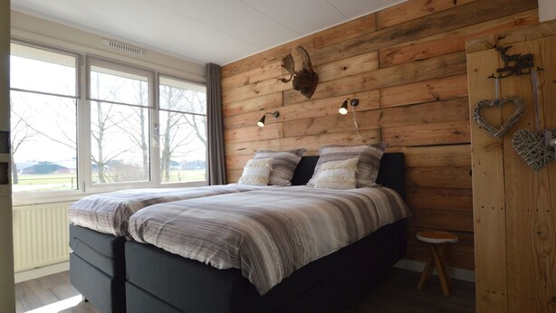 A bedroom with a large bed, wooden wall paneling, a small stool, and a window with a view of the outdoors. - Cozy Holiday Home in Nistelrode With Garden (Nistelrode)