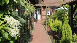 Um caminho de tijolos leva a uma casa de madeira com telhado de telhas, cercada por vegetação e plantas floridas.