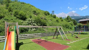 A playground with a slide, swings, and a red play area, set against a backdrop of green hills and mountains. - Mountain View Apartment in Going am Wilden Kaiser near Ski Area (Going am Wilden Kaiser)