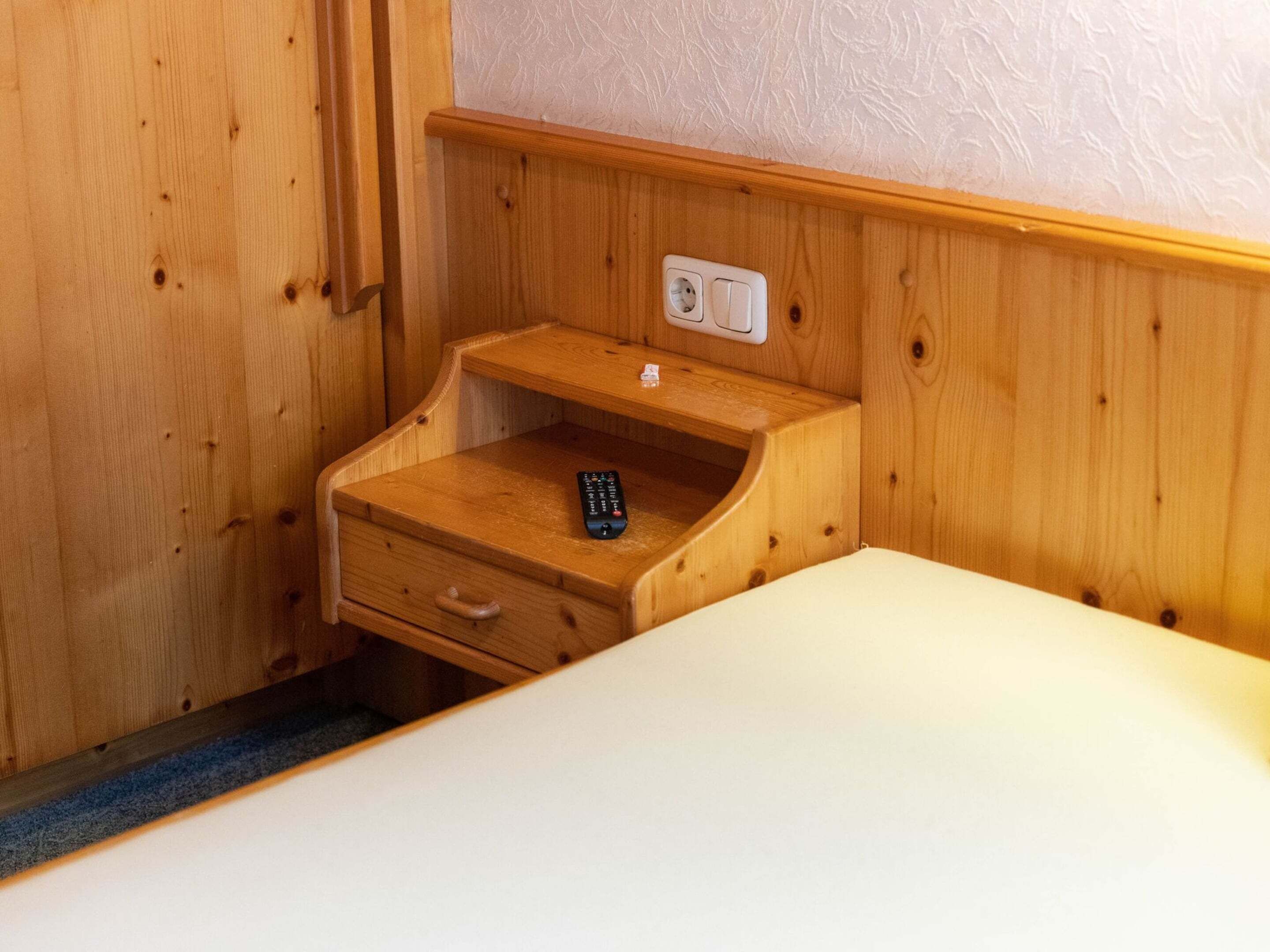 A wooden cabinet with a drawer and a shelf, a remote control on the shelf, and a power outlet on the wall.
