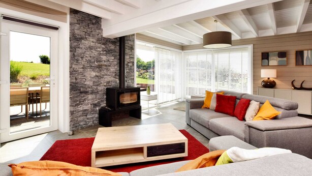 A modern living room with a stone fireplace, a grey sofa with colorful cushions, a wooden coffee table, and large windows offering a view of the outdoors. - Stylish Holiday Home in Houffalize With Sauna, Patio, Garden (Houffalize)