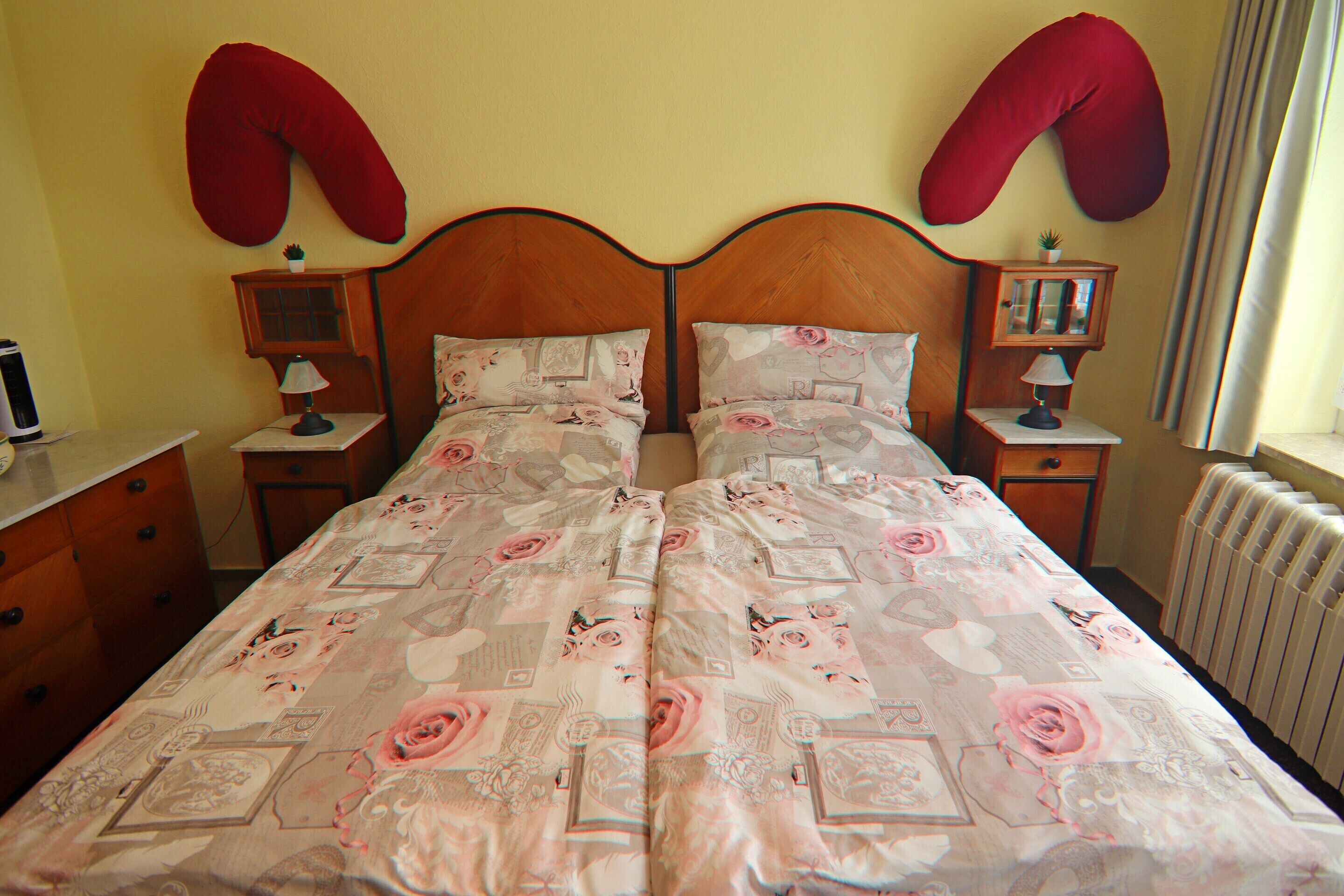 A double bed with floral bedding, two bedside tables with lamps, a red pillow on each side, and a radiator on the wall.