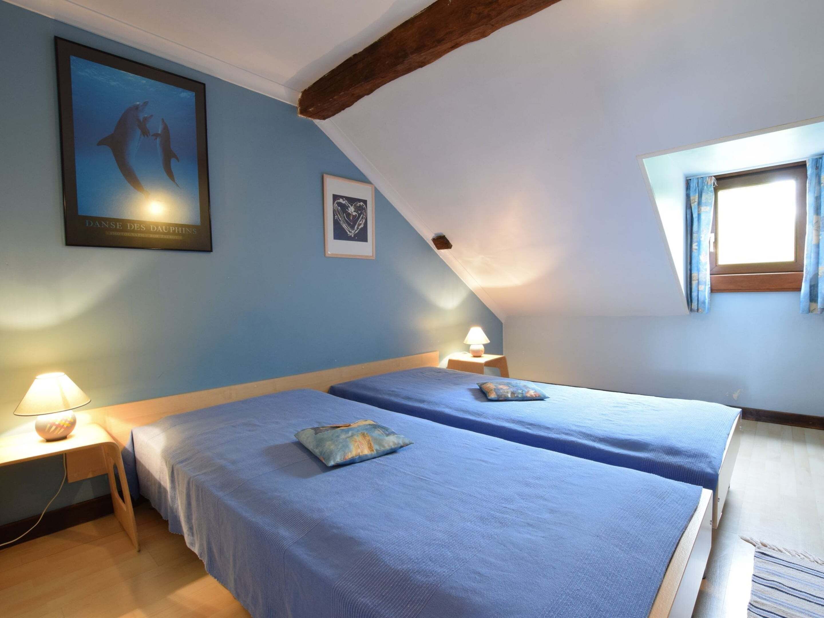 Een kamer met twee bedden, een ingelijst plaatje van dolfijnen, een tafeltje met een lamp en een raam met gordijnen.