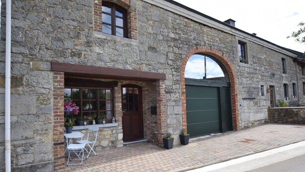 A stone building with a green garage door, a small outdoor seating area with chairs and potted plants, and a brick-paved sidewalk. - Cozy Family Stay in Durbuy (Durbuy)