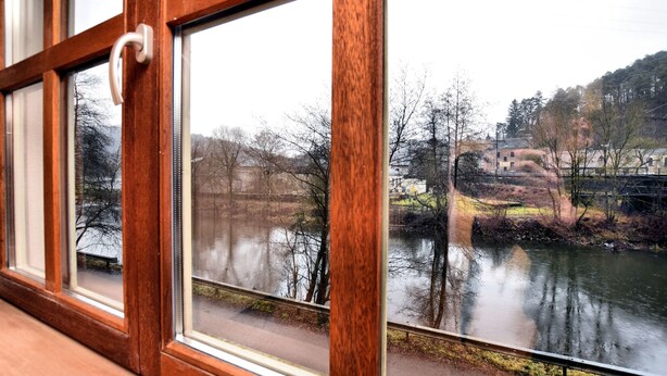 A window with a wooden frame and a white handle, overlooking a body of water with reflections of trees and buildings.