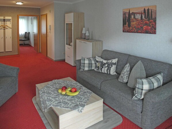 A living room with a grey sofa, a coffee table with fruit, a red carpet, a wardrobe, and a painting on the wall. - Ferienwohnung in der Nahe des Skigebietes (Willingen)