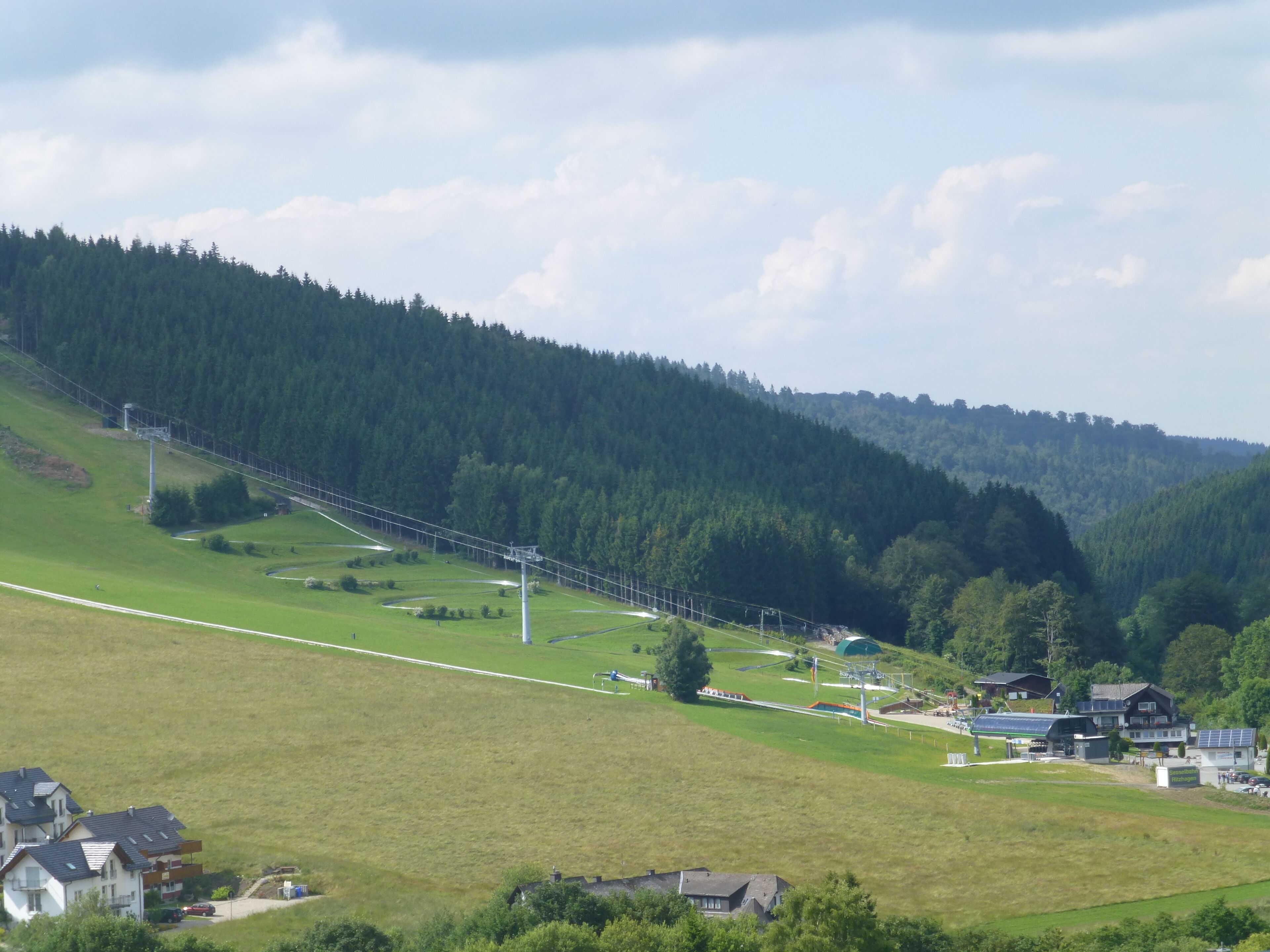 Paisagem rural com um teleférico, casas e uma colina arborizada.