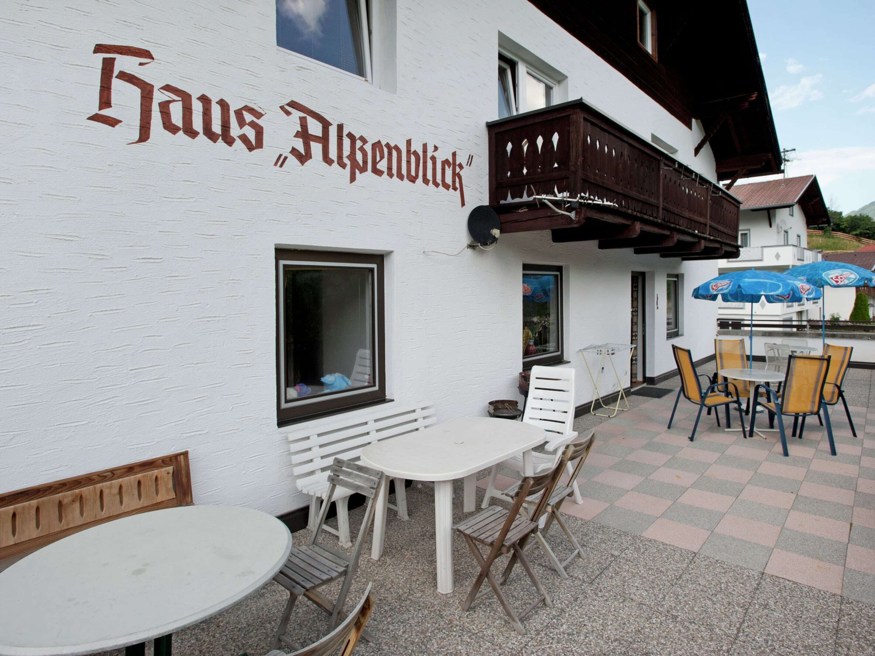 A building with a sign reading "Haus Alpenblick". There are outdoor tables and chairs, a wooden bench, and a window with a view.