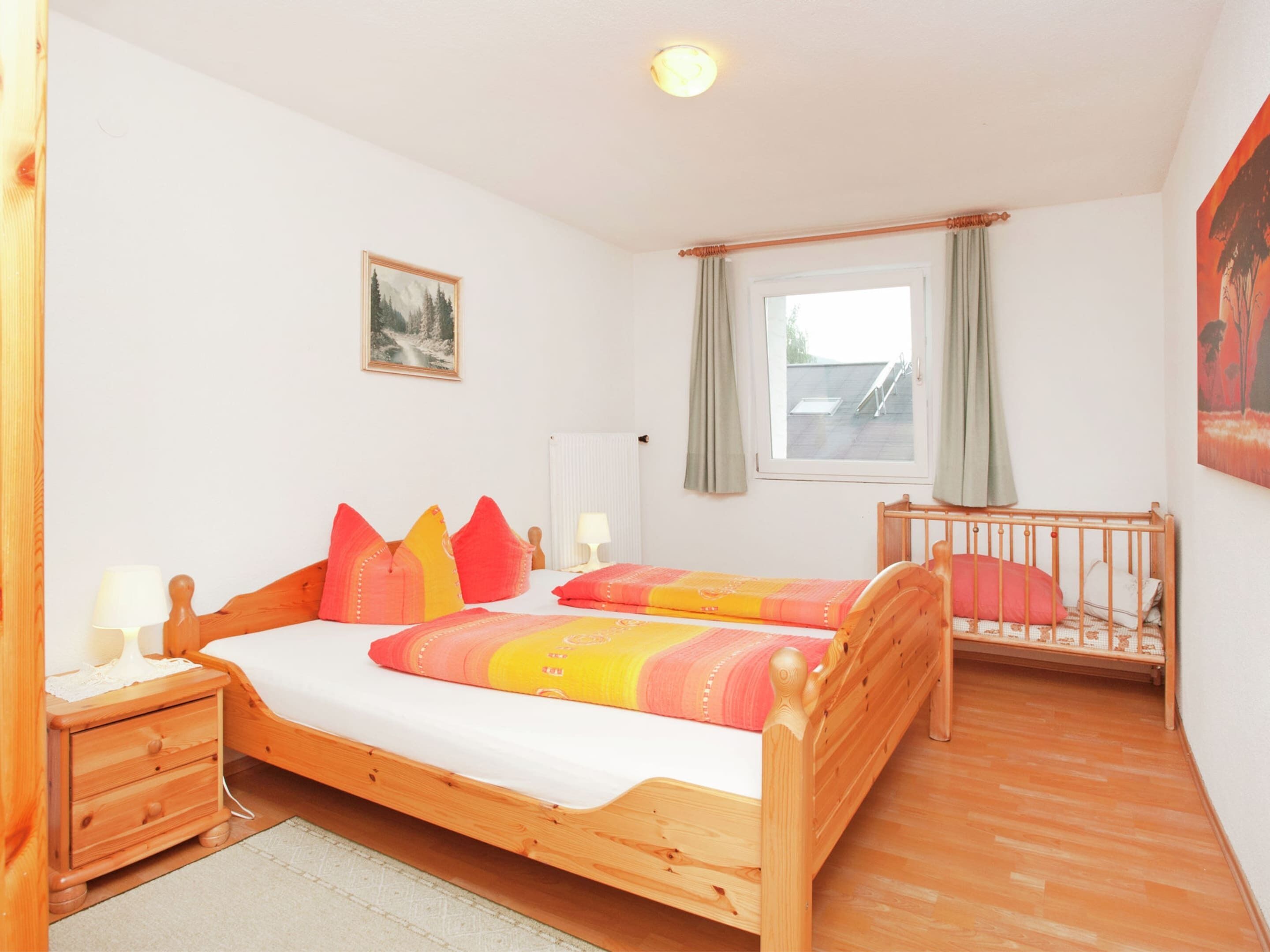 Ein Schlafzimmer mit zwei Holzbetten, einem Nachttisch, einem Fenster mit Vorhängen, einem Heizkörper und einem Bild an der Wand.