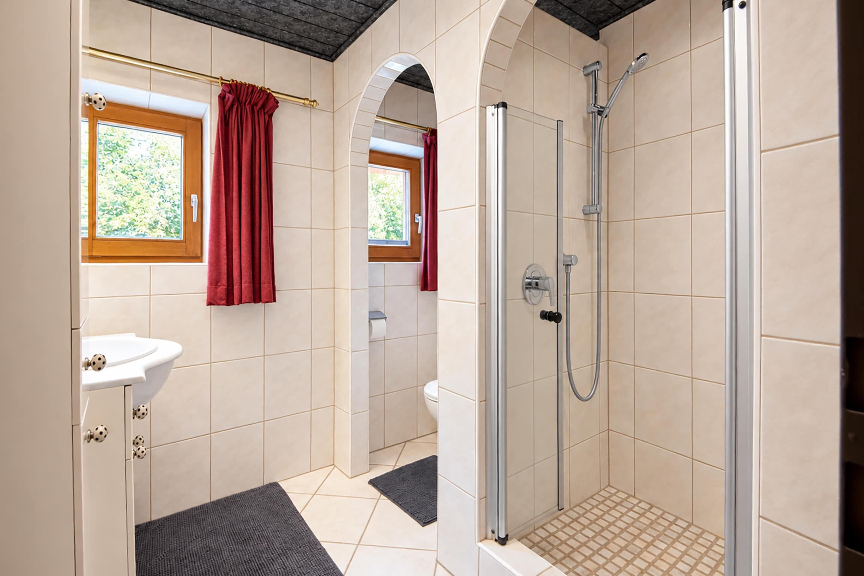 A bathroom with a shower, a sink, and a toilet. There are windows with curtains, and the floor has a patterned tile design.