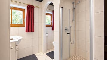A bathroom with a shower, a sink, and a toilet. There are windows with curtains, and the floor has a patterned tile design.