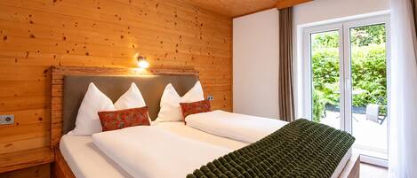 Dormitorio con paneles de madera, una cama, almohadas y una ventana con vista a follaje.