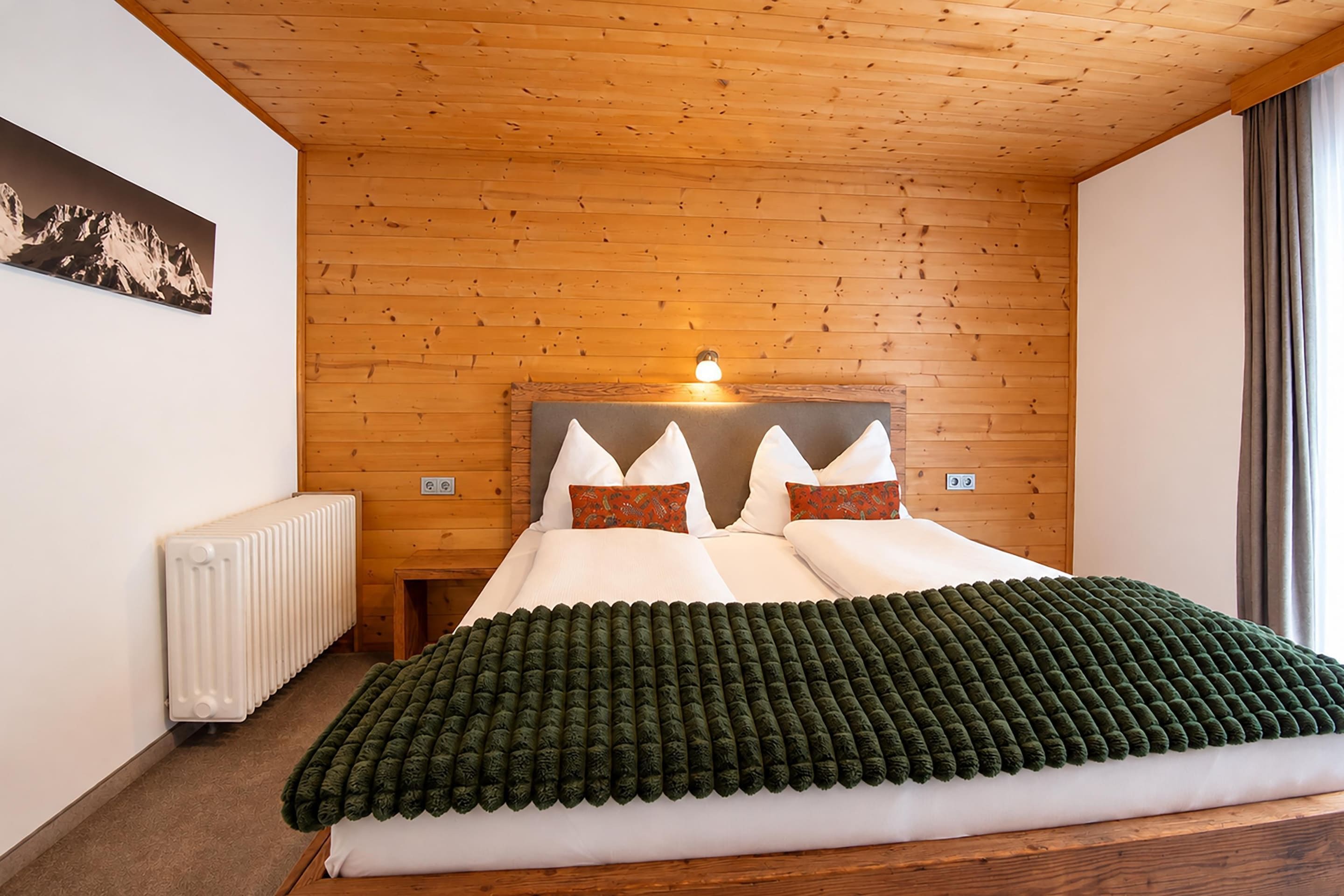 Une chambre avec un plafond en bois, un lit avec des oreillers blancs et orange, une couverture verte et un radiateur.