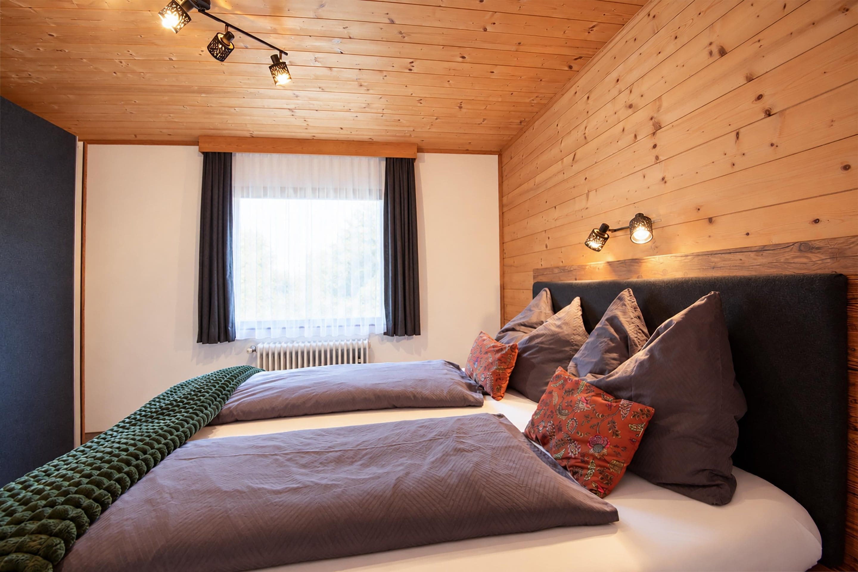 Una camera da letto con soffitto in legno, un letto con molti cuscini, una finestra con tende e un termosifone.
