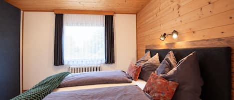 Una camera da letto con soffitto in legno, un letto con molti cuscini, una finestra con tende e un termosifone.