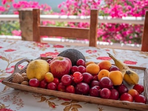 A tray of assorted fruits on a table with a floral tablecloth, set against a backdrop of pink flowers and a wooden chair.