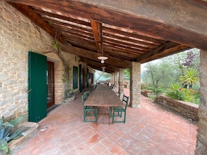 A covered outdoor dining area with a long wooden table and chairs, stone walls, and a view of greenery.