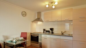 Een kleine keuken met een fornuis, oven en koffiezetapparaat, een eettafel met stoelen en een klok aan de muur.