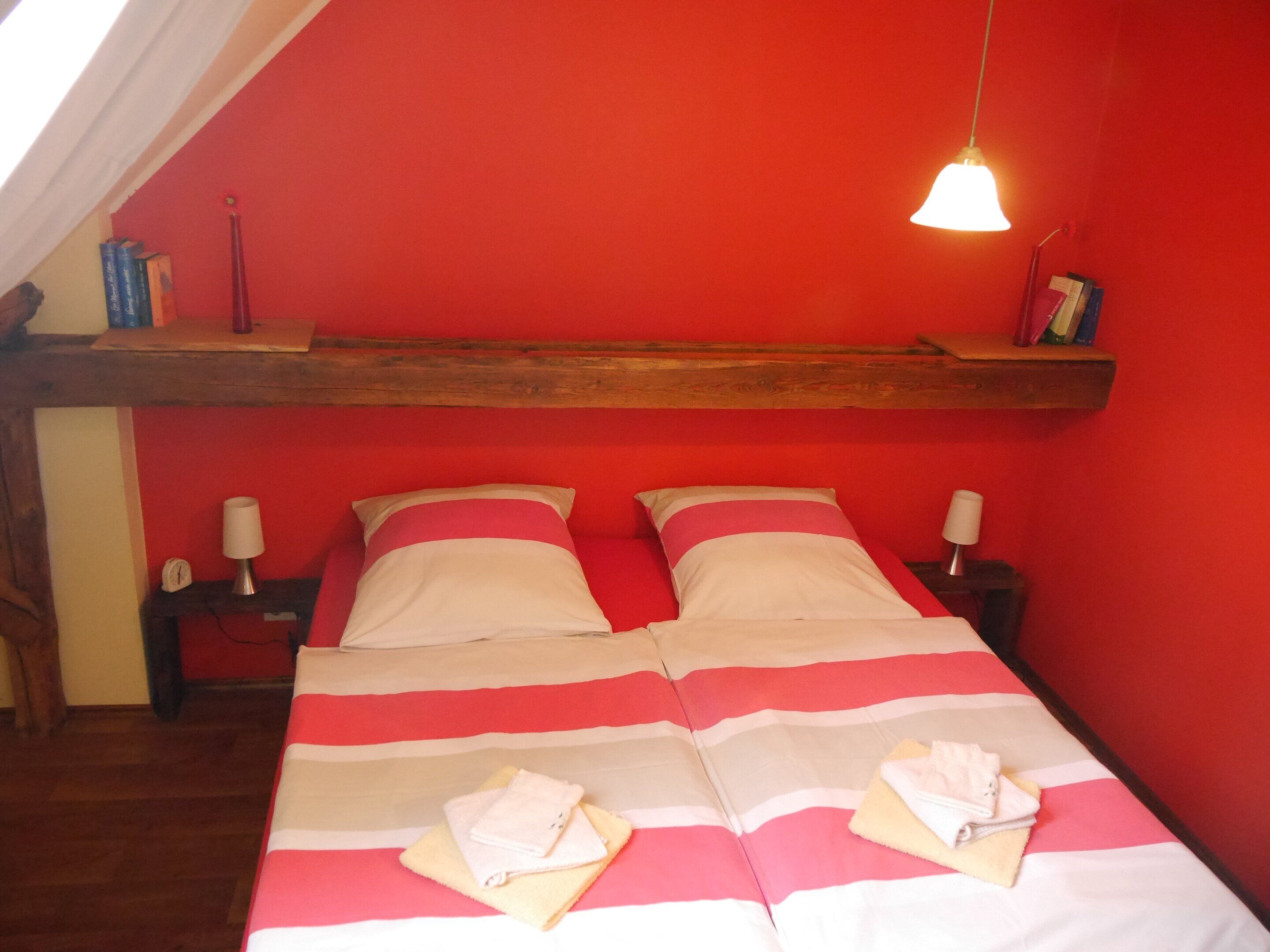 A room with a red wall, a wooden ceiling beam, a bed with striped bedding, two bedside lamps, and a hanging light fixture.