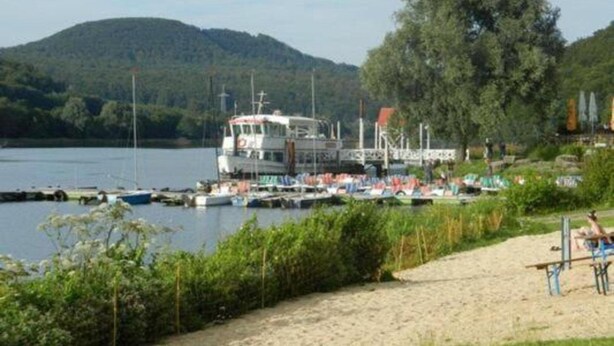 A ferry docked at a marina with boats, a sandy beach, and a forested hill in the background.