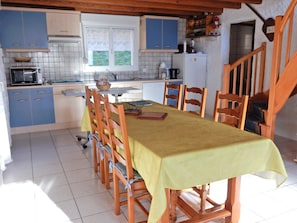 A dining area with a table set for four, wooden chairs, a kitchen in the background, and a window with lace curtains. - Holiday Home in Vosges With Garden and BBQ (Haut-du-Them-Château-Lambert)