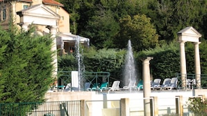 A swimming pool with a fountain, lounge chairs, and a building with columns in the background. - Apartment in Rhone Alpes Near Ski Slopes (Montbrun-les-Bains)