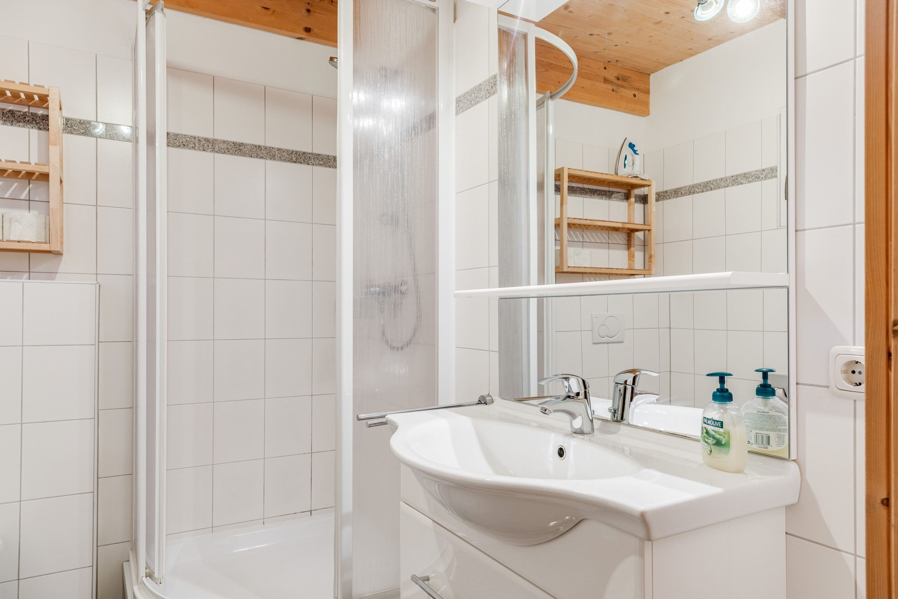 A bathroom with a shower, sink, and mirror. There are wooden shelves and a towel rack.