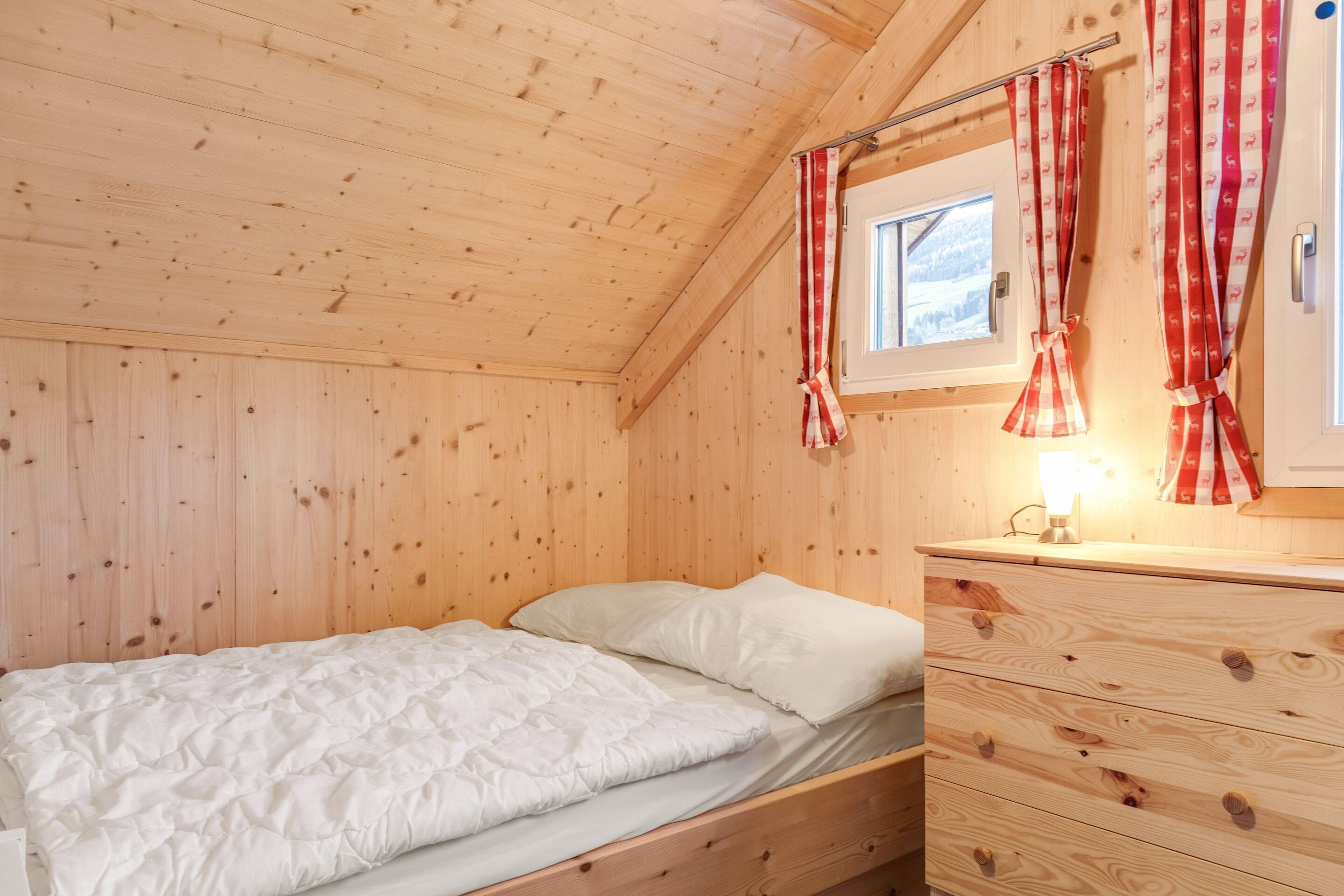 A wooden cabin room with a bed, a wooden dresser, and a window with curtains.