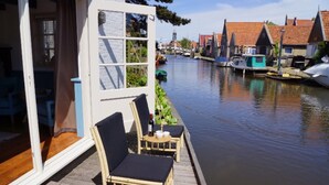 A wooden deck with two wicker chairs and a small table with a bottle and cups, overlooking a canal with boats and traditional buildings in the background. - Holiday Home in Hindeloopen by Lake (Hindeloopen)