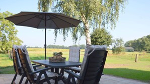 Um pátio externo com mesa, cadeiras, um guarda-sol e vista para um campo gramado.