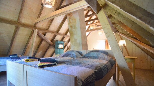 Quarto com teto inclinado, vigas de madeira, cama com colcha, mesa de cabeceira e luminária.