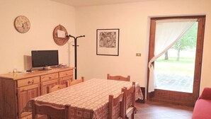 Uma sala de jantar com uma mesa posta para quatro pessoas, uma televisão sobre um móvel de madeira, um quadro na parede e uma janela com cortina.