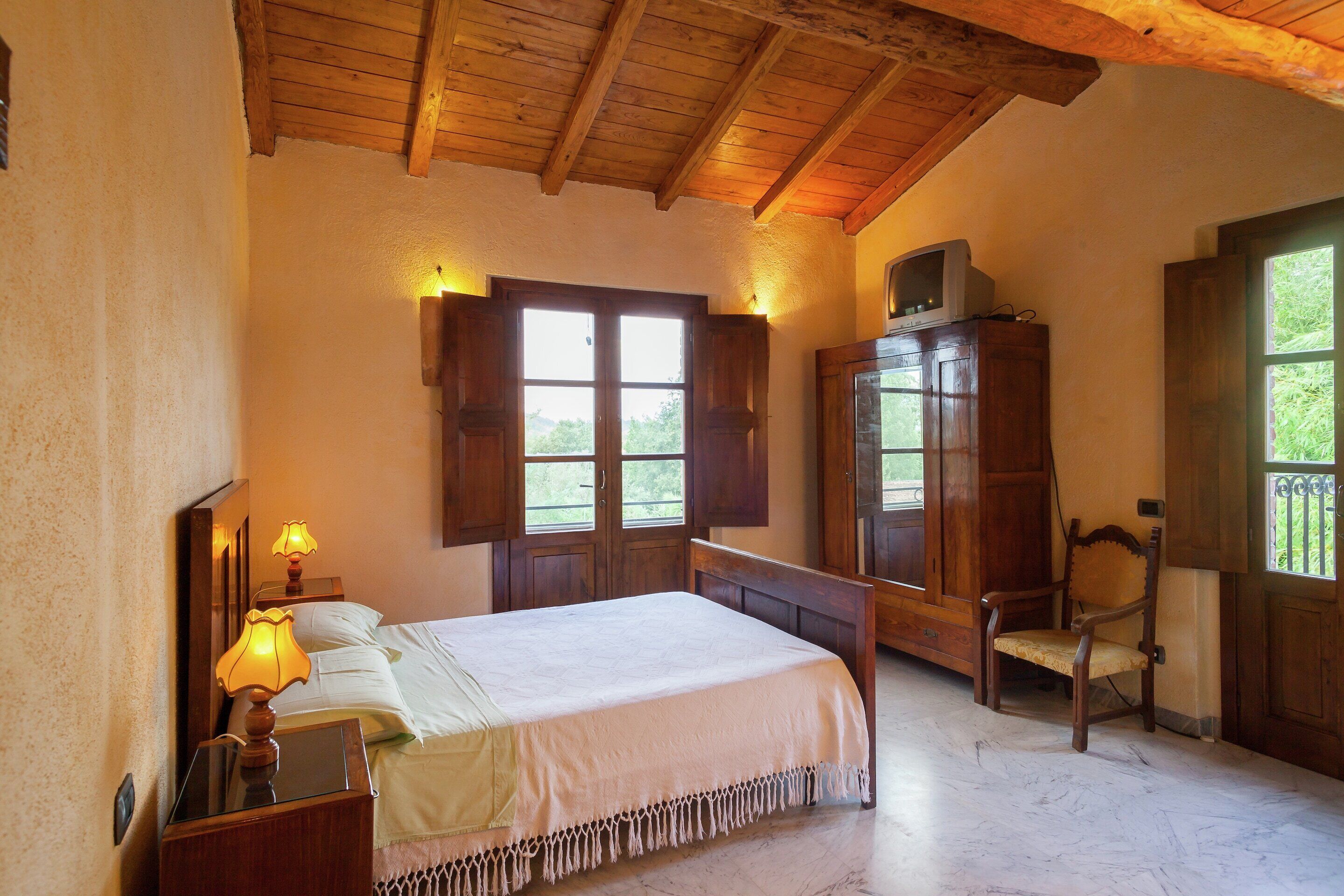 Un dormitorio con cama, cabecera de madera, mesita de noche con lámpara, armario de madera y ventana con contraventanas.
