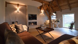 A cozy living room with a fireplace, wooden beams, a sofa, a coffee table, and a view of the outdoors.
