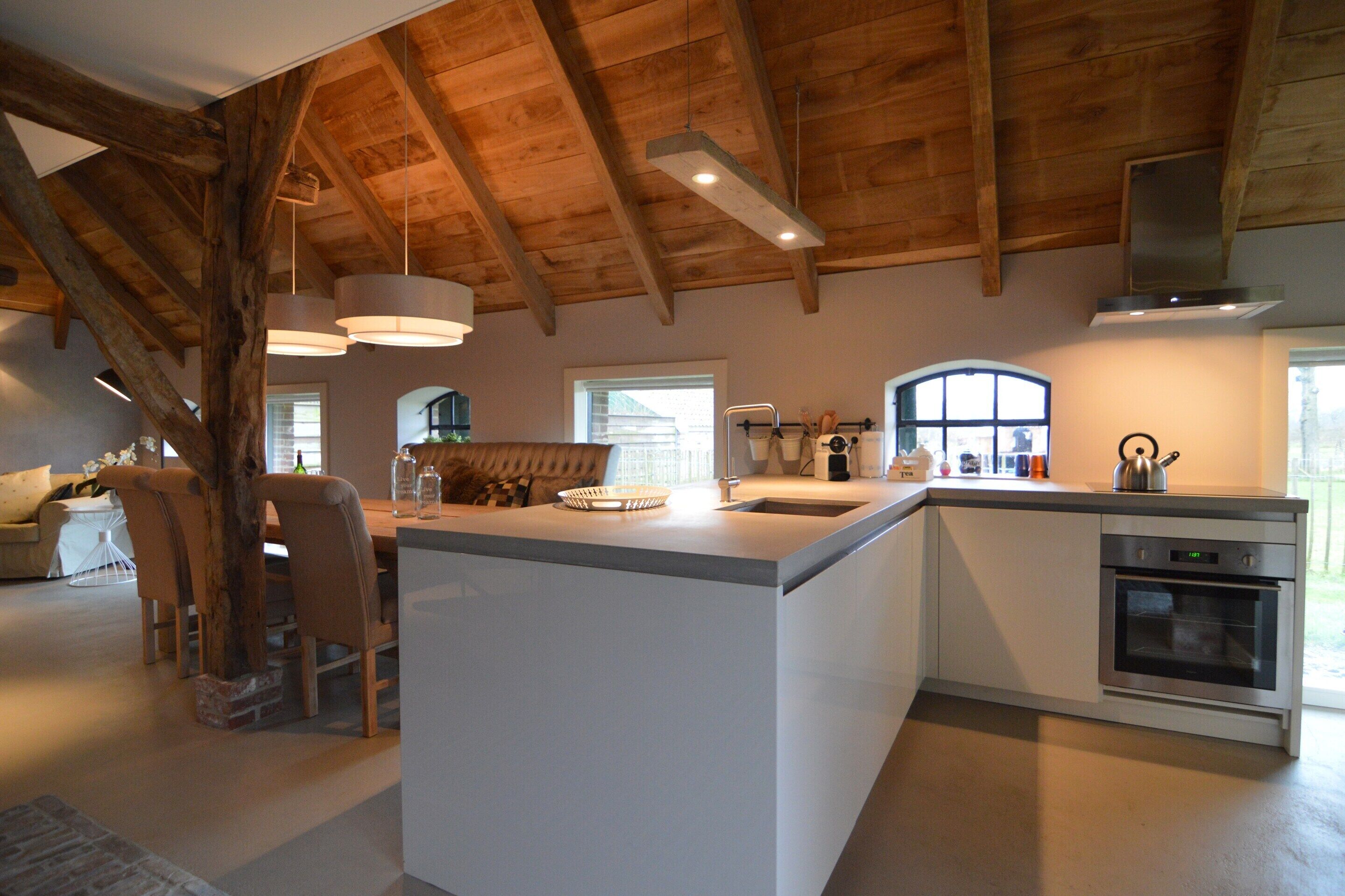 A modern kitchen with a large island, wooden ceiling beams, pendant lighting, and a view of the outdoors.