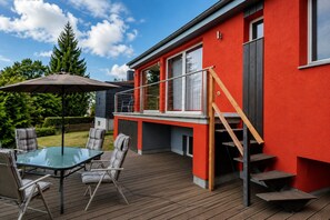 A modern outdoor deck with a glass table and chairs, a wooden staircase, and a red building facade. - Ski- und Wanderurlaub im Wald Ferienhaus Adlerblick (Masserberg)