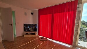 A room with a wooden floor, a television on a stand, and a red curtain. - Ski- und Wanderurlaub im Wald Ferienhaus Adlerblick (Masserberg)